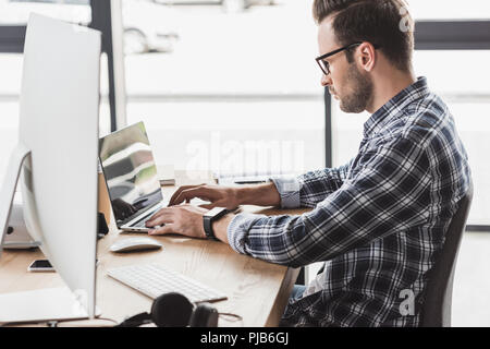 side view of handsome young programmer in eyeglasses working with laptop and desktop computer Stock Photo