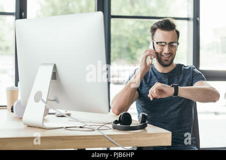 smiling young man in eyeglasses checking smartwatch and talking by smartphone at workplace Stock Photo