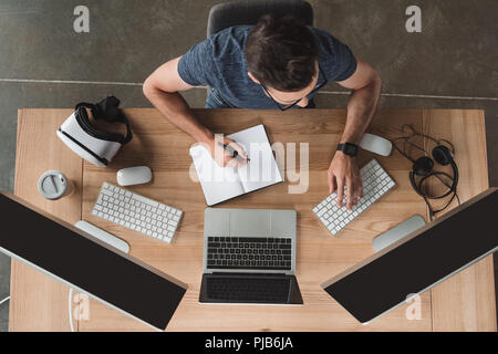 overhead view of young programmer taking notes in notebook and using computers at workplace Stock Photo