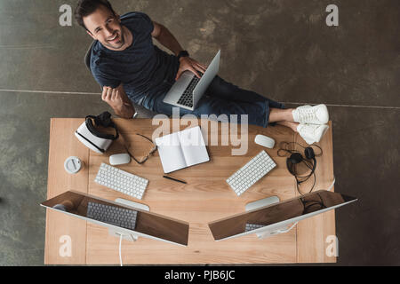 overhead view of handsome young programmer using laptop and smiling at camera Stock Photo