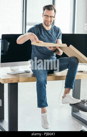 handsome smiling young programmer in eyeglasses eating pizza at workplace Stock Photo