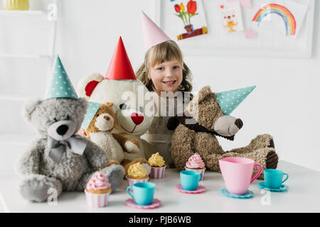smiling birthday kid with teddy bears in cones having tea party with cupcakes at table Stock Photo