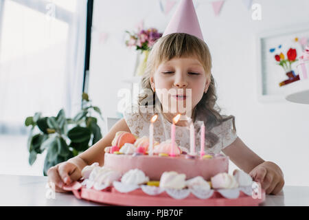happy adorable kid in cone blowing out candles from birthday cake Stock Photo