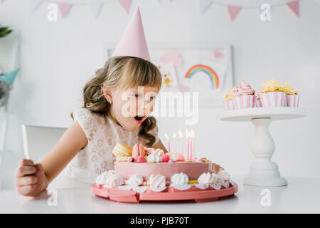 little birthday girl in cone blowing out candles from cake on table Stock Photo