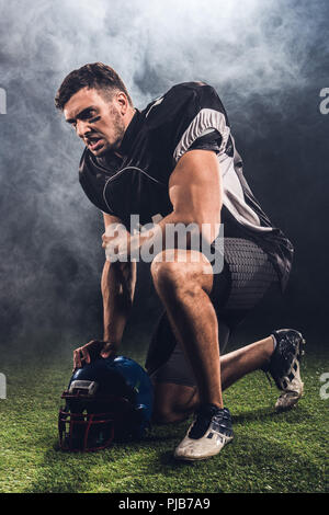 angry young american football player with helmet standing on knee on black Stock Photo