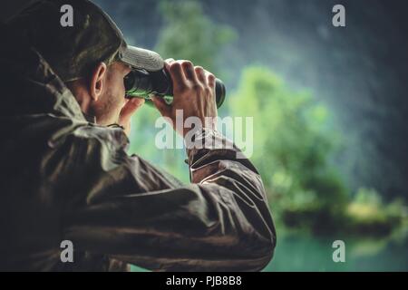 Caucasian Hunter in Masking Camouflage Uniform with Binoculars. Hunter Spotting Game. Poacher or Soldier Clothing. Stock Photo