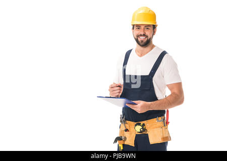 handsome happy workman in tool belt and hard hat writing on clipboard and smiling at camera isolated on white Stock Photo
