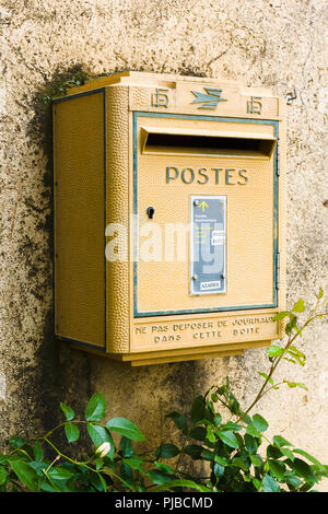 A classic vintage yellow French mailbox mounted on a textured stone wall, surrounded by green foliage at the bottom. Stock Photo