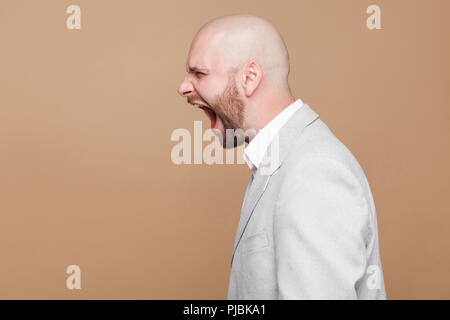 Profile side view portrait crazy anger of middle aged bald bearded businessman in light gray suit standing and screaming with oepn mouth. indoor studi Stock Photo