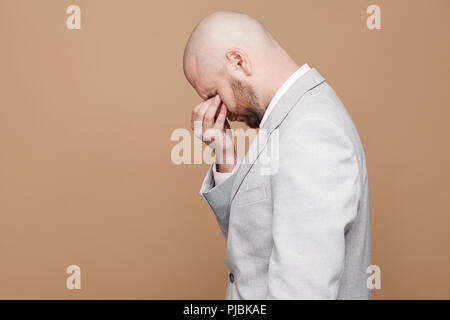 Profile side view portrait of sad crying middle aged bald bearded businessman in light gray suit standing and closed eyes with hands. indoor studio sh Stock Photo