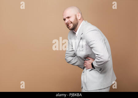 Stomach pain. profile side view portrait of middle aged bald bearded businessman in light gray suit standing and holding his painful belly. indoor stu Stock Photo