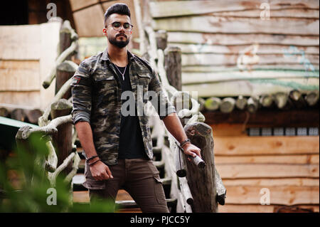 Awesome beautiful tall ararbian beard macho man in glasses and military jacket posed outdoor against wooden stairs. Stock Photo