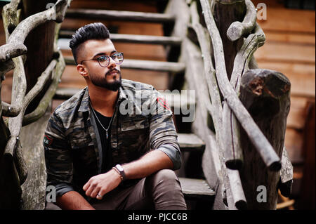 Awesome beautiful tall ararbian beard macho man in glasses and military jacket posed outdoor against wooden stairs. Stock Photo