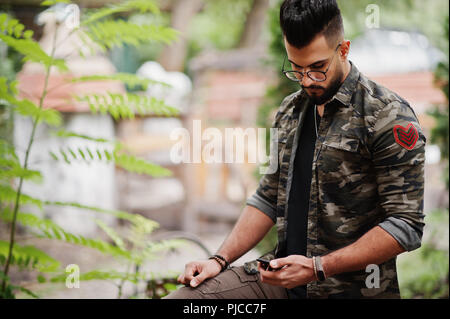 Awesome beautiful tall ararbian beard macho man in glasses and military jacket posed outdoor and looking at his mobile phone. Stock Photo