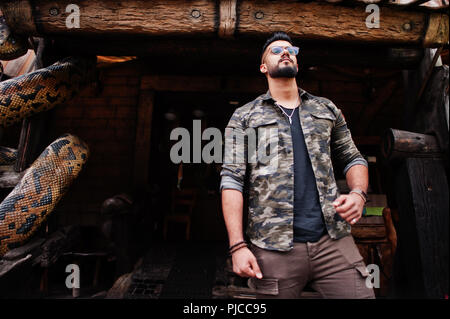 Awesome beautiful tall ararbian beard macho man in glasses and military jacket posed outdoor against snake python. Stock Photo