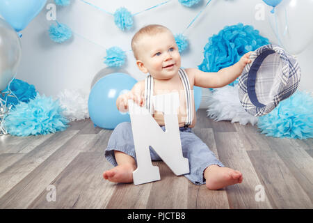 Portrait of cute adorable Caucasian baby boy with blue eyes barefoot  in pants with suspenders and hat, sitting on wooden floor in studio, holding lar Stock Photo