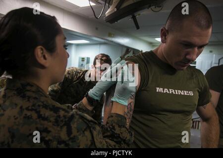 PACIFIC OCEAN- U.S. Navy Petty Officer 2nd Class Leslie Rodriguez, a hospital corpsman with the Command Element (CE), 13th Marine Expeditionary Unit (MEU), administers a smallpox vaccination to Gunnery Sgt. Skyler Tooker, communication strategy and operations chief with the CE, 13th MEU, aboard the Wasp-class amphibious assault ship USS Essex (LHD 2) during a regularly scheduled deployment of the Essex Amphibious Ready Group (ARG) and the 13th MEU, July 13, 2018. The Essex ARG/MEU team is a strong, flexible, responsive and consistent force capable of maneuver warfare across all domains; it is  Stock Photo