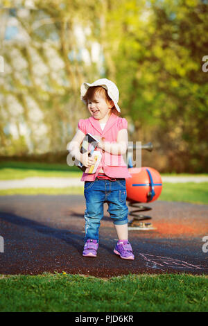 Colorful portrait of a cute smiling little girl toddler child, two or three years old, in pink jacket, blue jeans, white hat standing on playground ho Stock Photo
