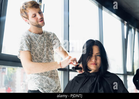 Close up. Female hairdresser straighten brown hair to woman using hair iron in beauty salon. Female stylist makes hairstyle for young beautiful woman in hairdressing salon. Stock Photo