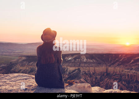 A view from the back of a girl in a hat sits on a mountain and admires the beautiful view and dawn or sunset in Cappadocia in Turkey. Stock Photo