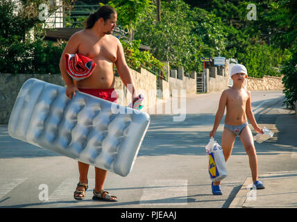 A man and boy walk down a street wearing beach attire carrying an inflatable floater and supplies for a day at the beach in Baska, Island Krk, Croatia Stock Photo
