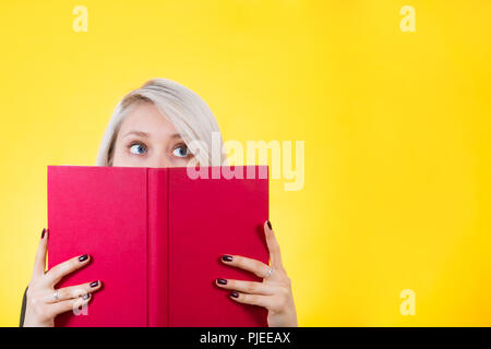 Dreamer girl hiding behind a opened red book looking away and imagine isolated over yellow background. Stock Photo