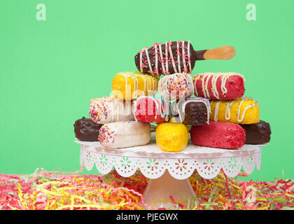 Close up on many colorful candy coated popsicle cake pops stacked on a white pedestal with bright paper party streamers below, bright green background Stock Photo