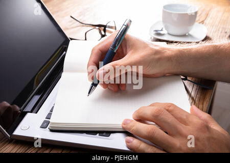 Person's Hand Writing On Black Notebook Using Pen Over Laptop Stock Photo