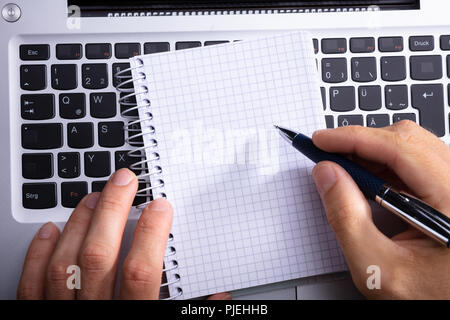 Person's Hand Writing On Spiral Notepad Over Laptop Keyboard Stock Photo