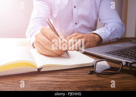 Person's Hand Writing On Black Notebook Using Pen Over Laptop Stock Photo