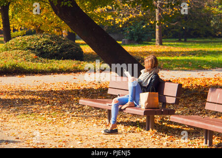 Woman is reading a book outside on the park bench, autumn leaves on the ground, beautiful autumn weather at city park covered by afternoon sun Stock Photo