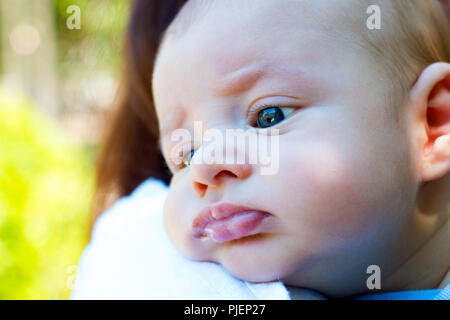 Cute little baby resting head on the mother arm, beautiful child with blue green eyes, close up portrait of the infant burping on mom's shoulder Stock Photo