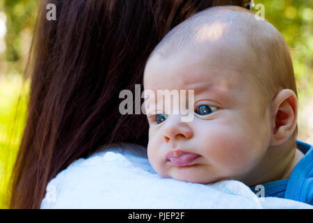 Portrait of beautiful baby in loving mother's arms, newborn head is resting on the shoulder to burp, cute infant face close up, green background Stock Photo