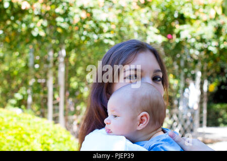 Little baby head from profile on the shoulder of young mother, pretty mom with make up on the eyes is holding baby, beautiful weather in park Stock Photo