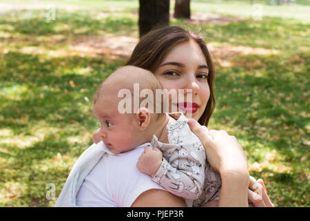 Beautiful mother holding her lovely baby outside in the park. Infant resting on the shoulder to burp while watching around, pretty happy mom smiling Stock Photo