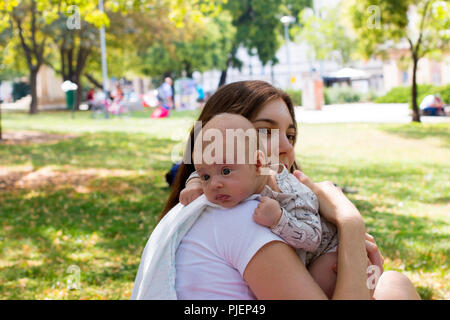 Portrait of beautiful baby resting head on mother arm, young mom is caring her infant in burping position after breastfeeding, outside in city park Stock Photo