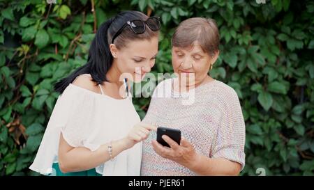 Granddaughter shows old grandmother something in smartphone, teaches her to handle with modern gadget and technology. Family, love, care concept Stock Photo