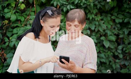Granddaughter shows old grandmother something in smartphone, teaches her to handle with modern gadget and technology. Family, love, care concept Stock Photo