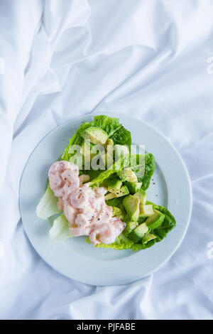 Classic prawn cocktail with pink marie rose sauce served with chopped avocado nestled in sweet cos lettuce cups on a white plate on crumpled white bed Stock Photo