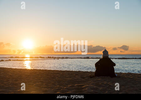 Happy woman sitting on cold beach with cup of tea with lights at sunset in Barcelona, Spain. Stock Photo