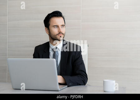 Handsome businessman is using a laptop, looking away and smiling while sitting on table in office. Stock Photo