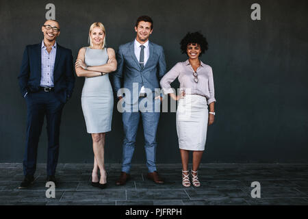 Full length portrait of successful business team standing together and smiling. Diverse business people posing for a photograph against dark wall. Stock Photo