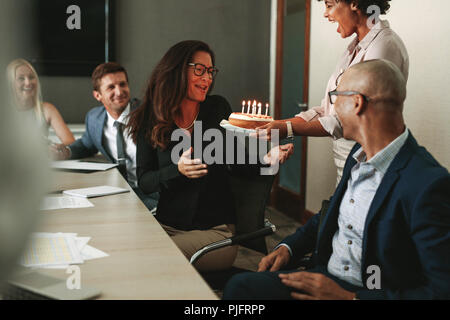 African woman giving a birthday cake to female executive in conference room. Workers celebrating colleague's birthday in office during a meeting. Stock Photo