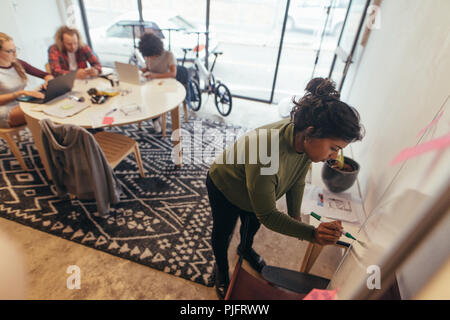 Young woman writing on whiteboard at meeting with colleagues working at the back. Young african female working at startup writing new project plan on  Stock Photo