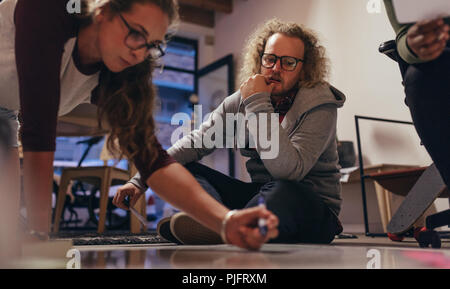 Team of tech professionals sitting on floor and brainstorming on new project plan. Woman writing project plan on board on floor with colleague looking Stock Photo