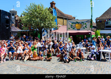 Crowd of people, audience, sitting at tables in cobbled town square in sunshine. Ground level view. Spectators at the Sandwich Folk and Ale festival Stock Photo