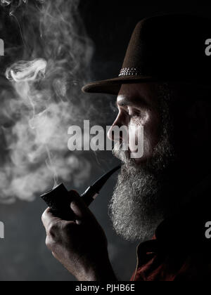 dramatic portrait of senior man in hat smoking tobacco pipe. Profile view of Austrian, Tyrolean, Bavarian old man Stock Photo