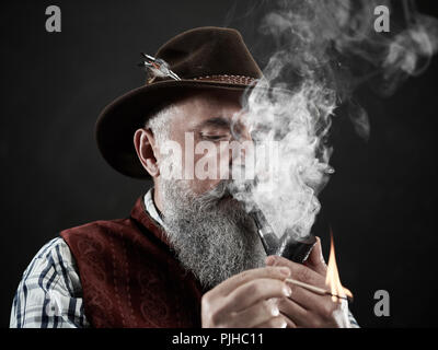 Black and white dramatic portrait of senior man in hat smoking tobacco pipe. Profile view of Austrian, Tyrolean, Bavarian old man Stock Photo