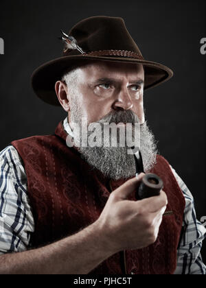 dramatic portrait of gray bearded senior man in hat smoking tobacco pipe. view of Austrian, Tyrolean, Bavarian old man in national traditional costume in retro style. Stock Photo