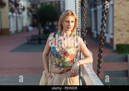 Beautiful young woman in tropical shirt, beige galligaskins with wicked bag standing with hand in pocket and posing on city background and looking at  Stock Photo